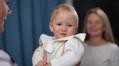Close-up portrait of curios baby girl looking back at blurred mother as doctor talking to adorable charming child. Caucasian pretty carefree toddler smiling on pediatrician visit in hospital