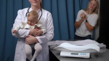 Front view dissatisfied infant in hands of unrecognizable doctor on the left with blurred mother talking at background. Unsatisfied Caucasian baby girl undergoing medical checkup in pediatric clinic