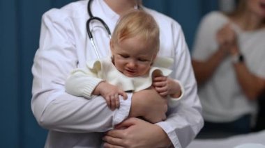 Portrait of dissatisfied baby crying in slow motion in hands of unrecognizable doctor shaking kid. Front view of unhappy little Caucasian toddler with sad facial expression on pediatrician visit