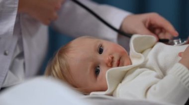 Close-up relaxed baby girl looking at camera and looking around as unrecognizable doctor listening kid with stethoscope and mother stroking child. Carefree toddler on visit in medical clinic