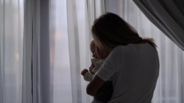 Silhouette of mother kissing baby girl in slow motion standing at window indoors. Back view medium shot of happy loving Caucasian woman enjoying morning with infant daughter