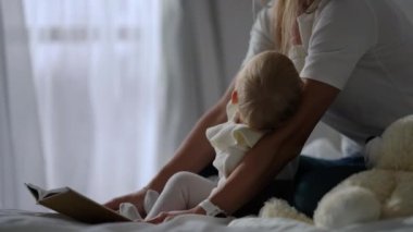 Curios little girl reading book with unrecognizable mother in slow motion sitting on bed at home. Side view portrait of happy relaxed confident Caucasian toddler enjoying hobby with parent