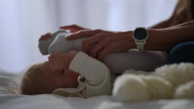 Charming baby girl lying on bed with mother hands bending kid in slow motion. Side view portrait of happy Caucasian infant playing with parent in bedroom at home