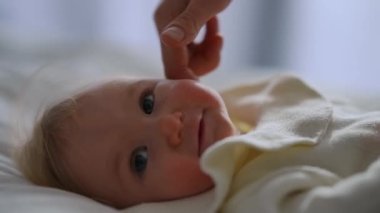 Headshot of cute infant looking around as female hand caressing face in slow motion. Unrecognizable Caucasian mother stroking baby girl cheek playing with calm adorable child lying on soft white bed