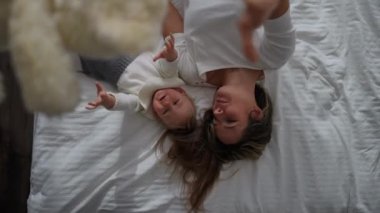 Top view laughing mother and baby daughter playing with teddy bear lying on white soft bed indoors. Cheerful Caucasian beautiful woman and excited pretty infant enjoying leisure in bedroom at home