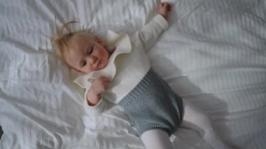 Top view portrait of relaxed baby girl lying on soft white cozy bed indoors looking around in slow motion. Happy carefree Caucasian infant toddler enjoying leisure in bedroom at home indoors