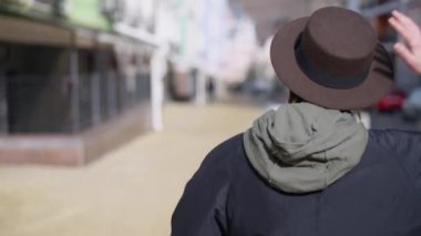 Back view joyful young attractive lady in hat strolling on city street turning looking at camera. Positive confident Caucasian woman walking on sunny day outdoors posing. Live camera tracking shot