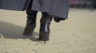 Back view female feet in high-heels walking on pavement on sunny autumn day outdoors. Unrecognizable confident elegant lady in boots and cloak strolling on city street. Confidence and femininity