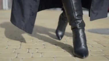 Front view steps of confident woman in black leather high-heel boots and cloak outdoors. Unrecognizable young stylish lady walking on pavement on city street on sunny day