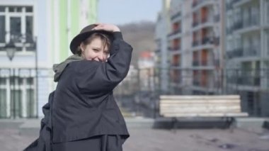 Live camera panning left to right as smiling young beautiful woman posing on city street in sunshine. Happy cheerful Caucasian confident female tourist resting in European town outdoors