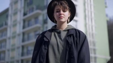 Pedestal shot of slim confident young woman in hat cloak and high-heel boots standing on city street. Beautiful Caucasian elegant lady posing on sunny day outdoors. Live camera moves down