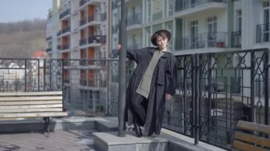 Young slim woman in cloak and high-heel boots standing at lamp post on city street looking away smiling. Positive confident Caucasian charming lady enjoying leisure outdoors