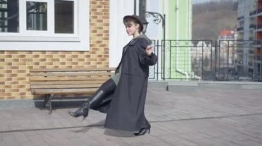 Funny carefree young woman in cloak and high-heels making steps looking at camera smiling. Wide shot side view of cheerful Caucasian slim lady in hat grimacing having fun on city street