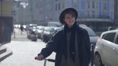 Zoom in to charming slim woman smiling looking around standing with kick scooter on city street in sunlight. Happy relaxed Caucasian beautiful lady in hat and cloak enjoying leisure outdoors in