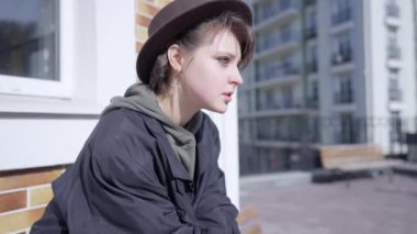 Side view sad young woman looking away thinking sitting on bench on city street. Beautiful slim Caucasian lady in hat procrastinating outdoors. Frustration and lifestyle concept