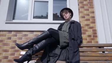 Wide shot confident woman posing in slow motion sitting on bench outdoors. Portrait of Caucasian stylish lady in high-heels cloak and hat looking at camera. Style and lifestyle concept