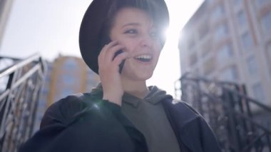 Joyful young woman with toothy smile picking up phone in slow motion talking laughing. Portrait of cheerful beautiful Caucasian lady in hat chatting standing in sunshine on city street