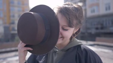Joyful brunette young woman with short hair laughing hiding with hat outdoors. Portrait of happy cheerful Caucasian lady looking at camera posing in slow motion in sunshine on city street