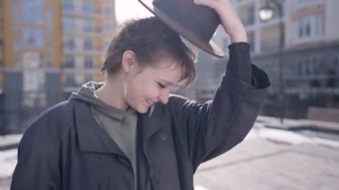 Woman putting on hat looking at camera smiling in slow motion standing on city street. Portrait of confident happy Caucasian elegant lady posing outdoors in sunshine