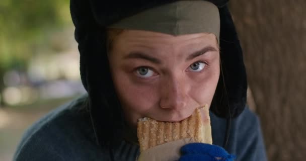 特写镜头 饥饿的无家可归的年轻女人慢吞吞地吃着面包 用灰色的眼睛看着相机 在夏季公园里拍摄的饥饿的可怜的高加索乞丐的头像 Cinema Prores — 图库视频影像