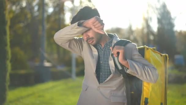疲惫的男人 提着食物袋站在阳光下 用夹克袖子擦拭额头 疲惫不堪的中东送货员站在城市公园里远眺沉思的画像 — 图库视频影像