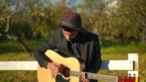 才能ある中東の男性ミュージシャンは 夏の公園で木製の柵にもたれてギターの歌を演奏します 太陽の下で屋外で趣味を楽しむスーツと帽子の熟練したハンサムな若い男の肖像画 — ストック動画