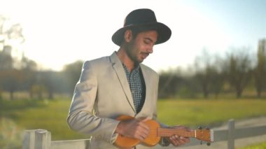 Şapkalı ve takım elbiseli, açık havada güneşli parkta ukulele çalan mutlu Orta Doğulu adam. Kendine güvenen genç müzisyenin portresi güneş ışığında hobisinin keyfini çıkarıyor.