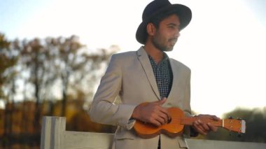 Ukulele çalan genç, zarif bir adam gün ışığında dışarıda durup ağır çekimde şarkı söylüyor. Parkta günbatımında müziğin keyfini çıkaran şapkalı ve takım elbiseli yetenekli Ortadoğulu müzisyenin portresi