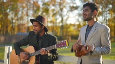 Gün batımında parkta gitar çalan ve ukulele çalan kendine güvenen iki zarif adam. İki yetenekli Orta Doğulu müzisyen açık havada gülümseyerek müzik dinliyorlar.