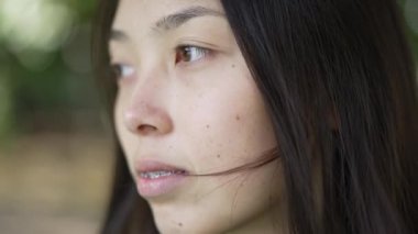 Siyah saçlı, kahverengi gözlü, düşünceli Asyalı genç bir kadının kameraya bakarken yüzünü çevirdiği bir fotoğraf. Açık havada yavaş çekimde poz veren çekici, zayıf bir kadının yakın plan portresi.