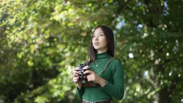 夏の公園でカメラと一緒に立って自信を持って才能のあるアジアの女性写真家の肖像画離れて見て笑っている 晴れた日には屋外で趣味を楽しむ若いスリム女性の中出し スローモーション — ストック動画