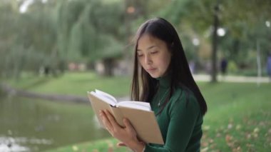 Parktaki çimlerin üzerinde oturmuş kitap okuyan gülümseyen kadın portresi. Güneşli bir günde açık havada hobi yapan güzel, ince Asyalı bayan. Bireysellik ve zeka kavramı