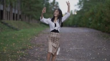 Sonbahar parkında canlı yayın yapmak için akıllı telefon kamerasıyla konuşan kendine güvenen neşeli Asyalı blog yazarı portresi. Genç, sıska, güzel bir kadın dışarıda blog yazıyor, gülümsüyor. Yavaş çekim