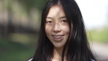 Genç gülümseyen Asyalı kadın portresi diş telleriyle kameraya bakıyor yavaş çekimde saça dokunuyor. Sunny Park 'ta poz veren kendine güvenen çekici bir kadının ön plan fotoğrafı.