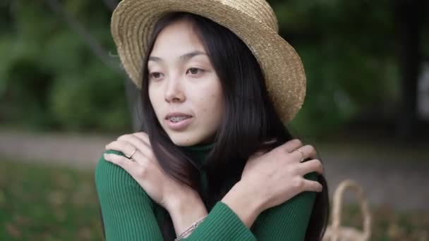 迷人的亚洲年轻女子微笑着拥抱着自己环顾四周 穿着草帽的美丽纤细女士的画像 在夏日公园的室外享受休闲 慢动作 — 图库视频影像