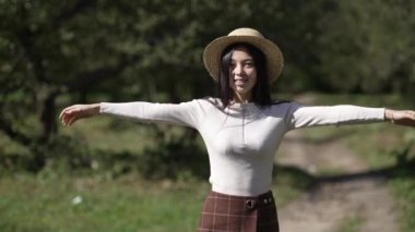 Hasır şapkalı gülümseyen kadın güneşli parkta ağır çekimde yürüyen kameraya bakıyor. Kendine güvenen neşeli Asyalı kadının portresi. Gün ışığında dışarıda geziniyor.