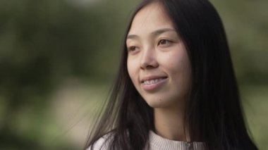 Rüzgârla gülümseyen, uzun siyah saçları ağır çekimde savuran çekici bir kadın. Tatmin olmuş Asyalı bir kadının portresi. Hafta sonu parkta eğleniyor.