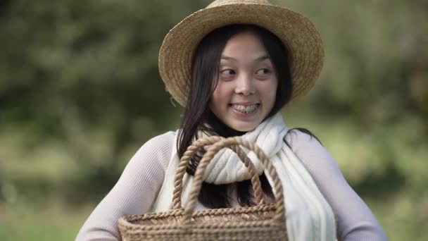 公園に立って屋外のわら籠でポーズ楽しい千年紀の女性の肖像画 幸せな自信に満ちたスリム美しいですアジアの女性でわら帽子とともに歯のブレース見ますカメラ笑顔 — ストック動画