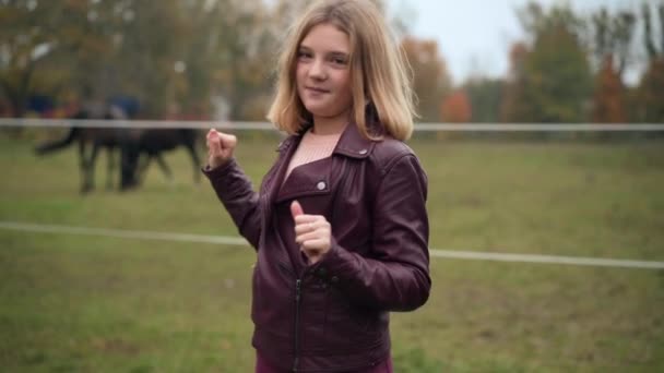 背景に馬の放牧を指してカメラを見て自信を持ってティーンの女の子 魅力的な白人10代のブロガーの肖像 秋の公園で牧草地にポーズ優雅なStalllionについて語る — ストック動画
