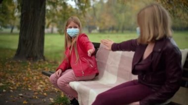 Covid maskeli şirin bir kız. Arkadaşının elini uzatıyor. Bankta oturuyor ve sosyal mesafeyi koruyor. Beyazların sonbahar parkında Coronavirüs salgınında buluşması