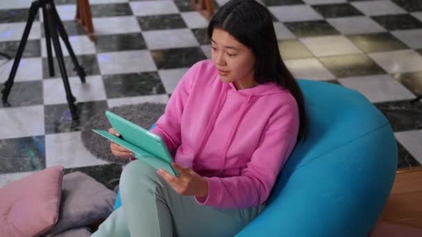 幸せなアジアの10代の女の子の高角度ビューの肖像画は笑顔を見上げデジタルタブレットを抱いている 成功10代のブロガー満足結果で座っています上のバッグ椅子でリビングルームに自宅 — ストック動画