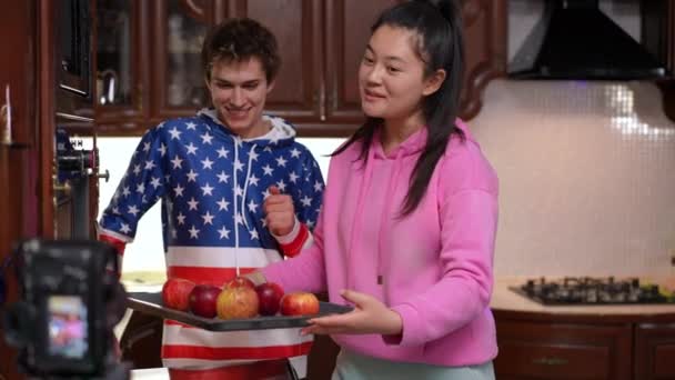 正のブロガーは 5をジェスチャーオーブンでリンゴとトレイを入れて話しています 幸せな自信に満ちたアジアの女の子と白人の男の子ライブストリーミング調理おいしいヘルシーなデザートでキッチン屋内 — ストック動画