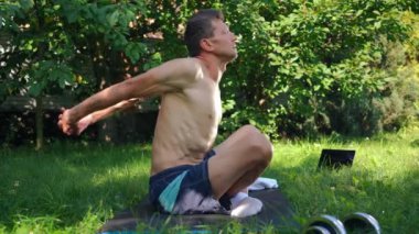 Yandan bakan adam nilüfer pozisyonunda oturarak ısınıyor ve gözleri kapalı meditasyon yapıyor. Güneşli bir yaz gününde arka bahçede, kendinden emin, Kafkas sporcusu eğitiminin geniş açılı portresi.