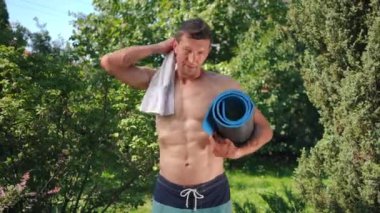 Omzunda havluyla dışarıda egzersiz paspasıyla poz veren kendinden emin sporcu. Yaz günü arka bahçede kameraya bakan beyaz bir adamın orta boy portresi.