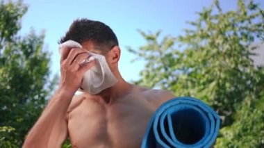 Beyaz tenli sporcu, terini siliyor ve dışarıda spor paspasıyla gülümseyen kameraya bakıyor. Mavi gökyüzü ve yeşil ağaçların arkasında poz veren kendinden emin bir adamın portresi