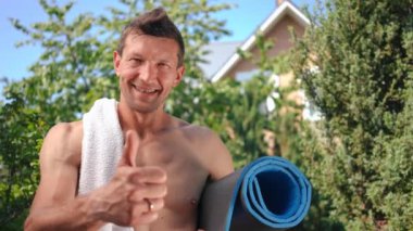 Mutlu beyaz adam başparmağını kaldırmış, gülümsüyor arka bahçede spor paspası ve havluyla kameraya bakıyor. Dışarda poz veren pozitif memnun sporcunun portresi