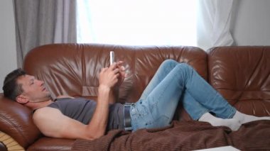 Tasasız Kafkasyalı bir adam kanepeye uzanmış akıllı telefon uygulamasında sesli gülüyor. Haftasonu boş zamanlarında sosyal medyada gezinen rahat, pozitif adamın geniş açılı portresi