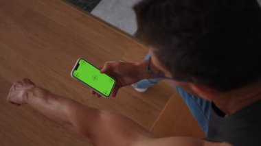 Yüksek açılı adam evde oturan yeşil ekran akıllı telefonu kullanıyor. Tanımlanamayan beyaz bir adamın omzunun üstünden çekim yapmak internette mobil uygulama kullanarak internette gezinmek.