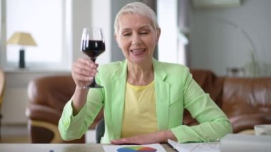 Neşeli orta yaşlı zarif kadın kameraya bakarak kadeh kaldırıyor camdan kırmızı şarap içiyor. Görüntülü sohbet POV 'uyla selamlaşan Kafkas iş kadınının ön manzara portresi. Yavaş çekim