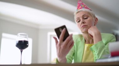 Parti şapkalı, üzgün, orta yaşlı bir kadın sağ telefon mesajlarını kontrol ediyor ve ağır çekimde bir bardak kırmızı şarap içiyor. Kafkasyalı, tatilde evde yapayalnız, üzgün bir beyaz portresi.